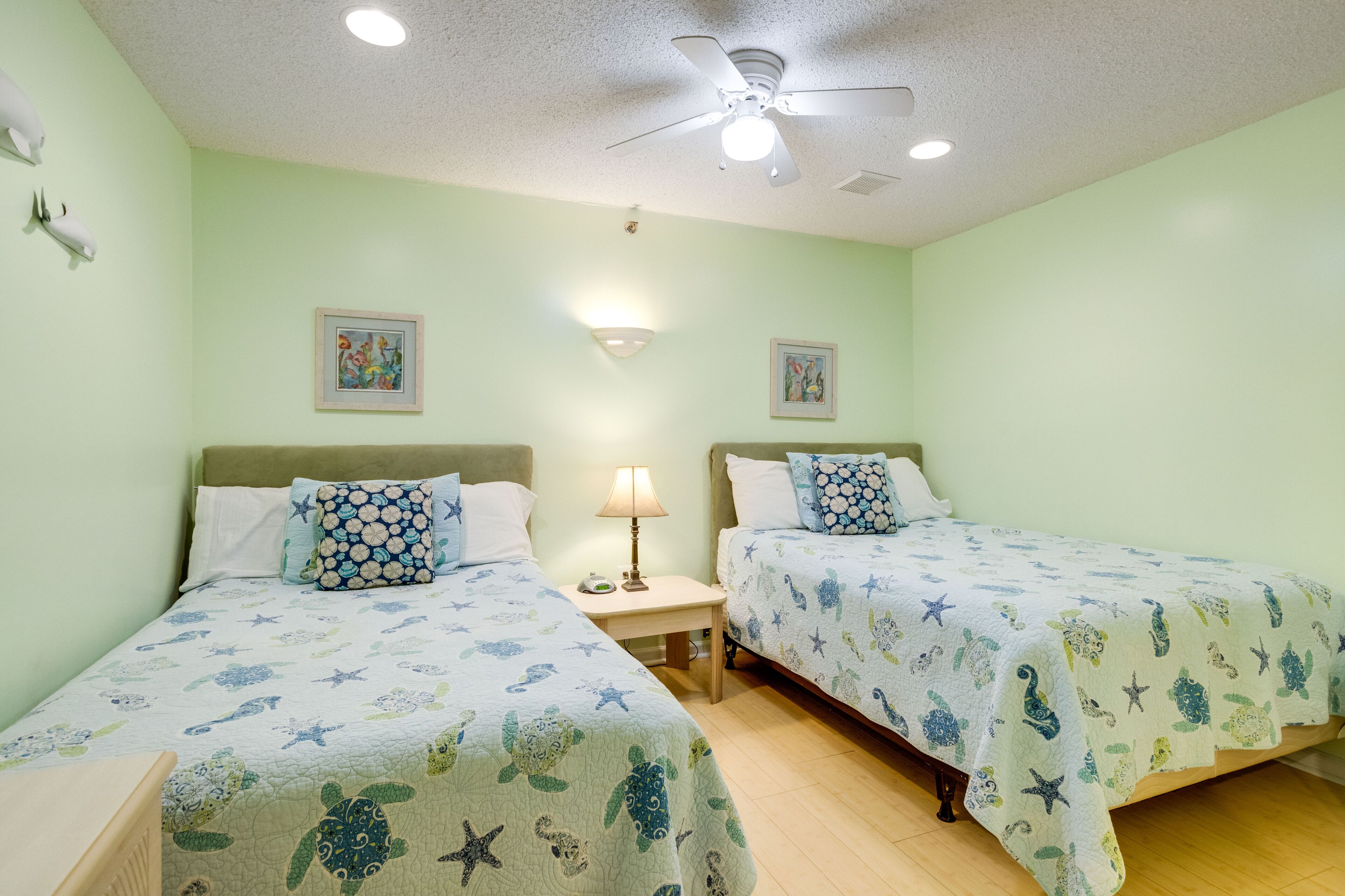 Une chambre avec deux lits, chacun agrémenté d’une couvre-lit sur le thème des étoiles de mer et des tortues, d’un ventilateur de plafond et de deux lampes fixées au mur.