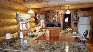 A log cabin kitchen with granite countertops, wooden cabinets, and wicker chairs. - Vermont Lake Front Log Cabin Close to Mt. Snow (Whitingham)