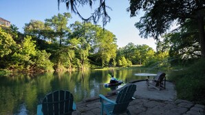 Un patio junto al lago con sillas Adirondack y una mesa, con vistas a un lago tranquilo rodeado de árboles.