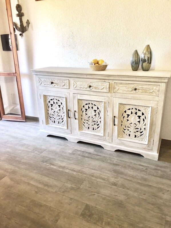 A white, intricately carved wooden cabinet with three drawers and two doors, placed against a textured wall.