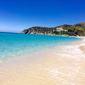 A beach with clear turquoise water, a sandy shore, and a building on a hillside.