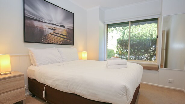 Una cama bien hecha con sábanas blancas, una lámpara de noche y una ventana con vistas al verdor.