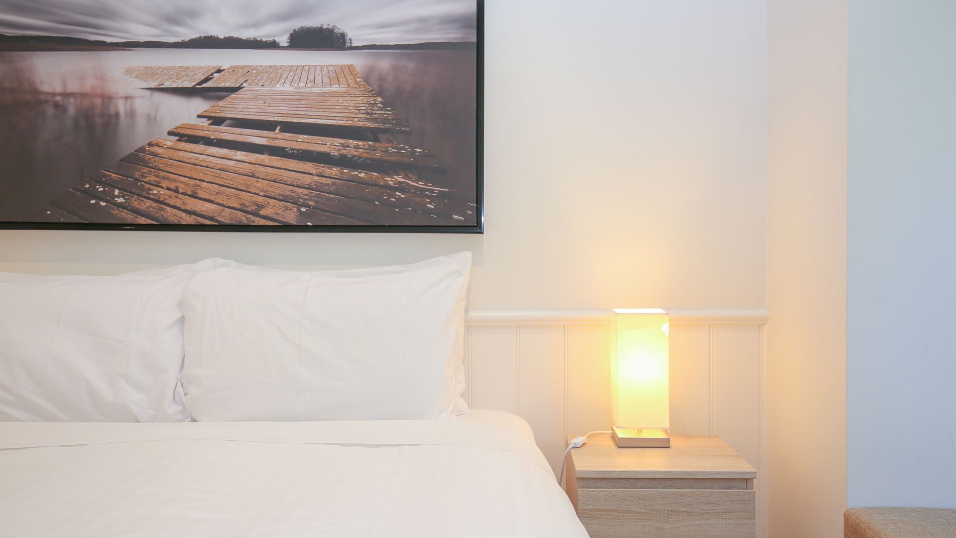 A hotel room with a bed, white pillows, a nightstand with a lamp, and a picture of a dock on the wall.