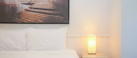 A hotel room with a bed, white pillows, a nightstand with a lamp, and a picture of a dock on the wall.