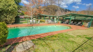 Una piscina rectangular con borde de ladrillo, rodeada de césped y un porche cubierto con sillas y sombrillas.