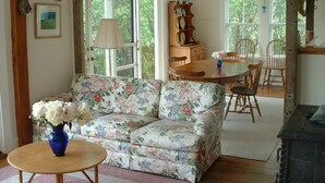 A living room with a floral sofa, a round wooden table, and a dining area with chairs and a wooden cabinet. - Charming Antique Cape, Water View, Historic Castine, Penobscot Bay. (Castine)
