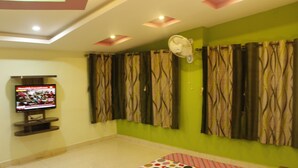 A room with a television on a stand, a bed with a polka dot bedspread, and curtains on the windows. - Hotel Radhakrishna (Wai)