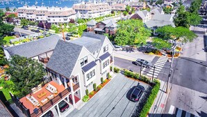 A residential area with a two-story house featuring a balcony, a driveway, and a street view with parked cars and a pedestrian crossing.