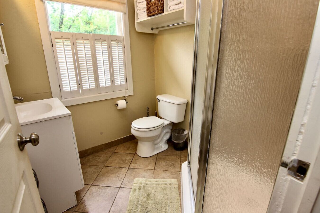 A small bathroom with a toilet, a sink, a window with shutters, and a towel rack.