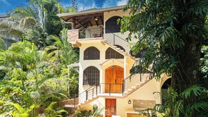 Ein mehrstöckiges Haus mit Treppe, umgeben von üppiger Vegetation und Palmen.