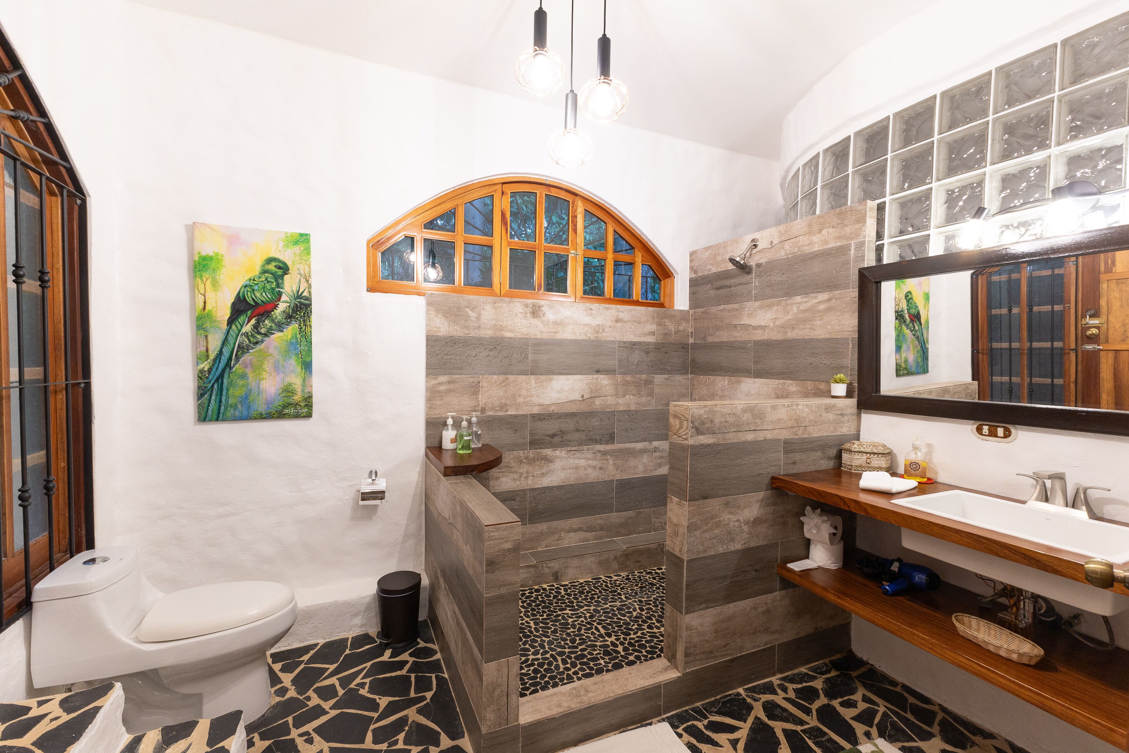 A bathroom with a white toilet, a shower with a wooden arch, a sink with a mirror, and a tiled floor.