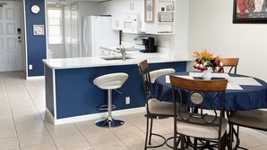 A modern kitchen with a central island, white cabinets, and a dining area with a blue tablecloth and two bar stools. - Fall Savings! Gulf-View 2BR2BA 
Beach Condo with private balcony. (Indian Rocks Beach)