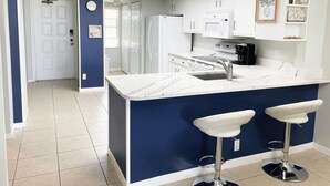 A modern kitchen with a central island, two bar stools, white cabinetry, and a decorative vase on the wall. - Fall Savings! Gulf-View 2BR2BA 
Beach Condo with private balcony. (Indian Rocks Beach)