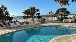 A pool with clear water, surrounded by a white fence and palm trees, with a view of the ocean in the background. - Fall Savings! Gulf-View 2BR2BA 
Beach Condo with private balcony. (Indian Rocks Beach)