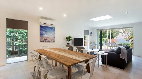 A modern living room with a wooden dining table, clear plastic chairs, a sofa, and a TV. - Depper St - Charming Beach Side Home in Lovely Location (SUNSHINE BEACH)
