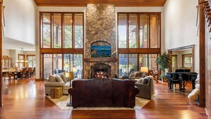 A spacious living room with a stone fireplace, large windows, and a piano. - CDAs Finest Secluded Waterfront Estate 7000 Sq ft 5 Forested Acres-Close to town (Post Falls)