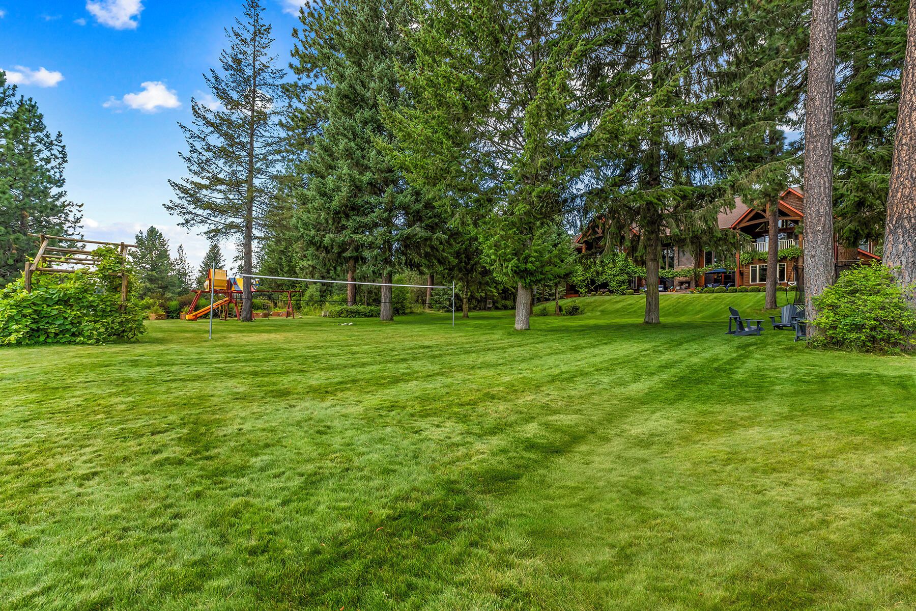 A well-maintained grassy area with a playground, tall trees, and a wooden building in the background.