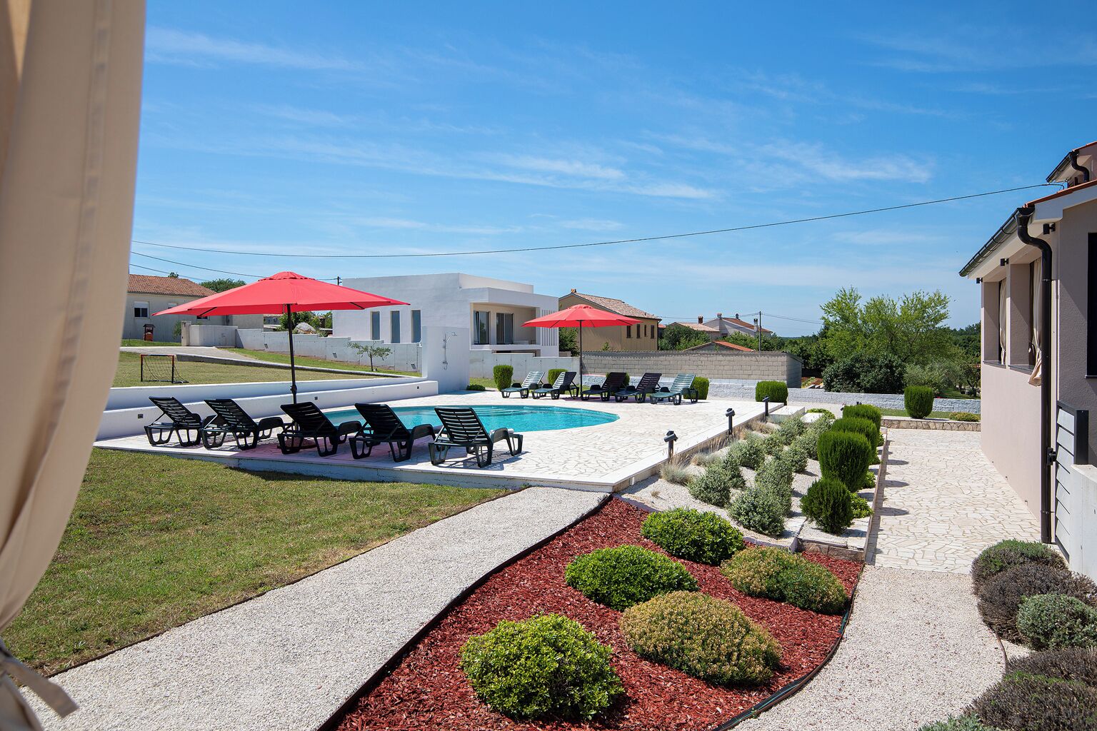 Un espace piscine agrémenté de chaises longues et de parasols rouges, entouré d’un aménagement paysager soigné et surplombé d’un ciel dégagé.