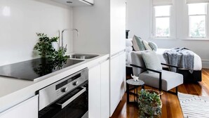 A modern kitchen with white cabinetry, a stainless steel oven, and a sink. There is a small round table with a plant beside the sofa. - Designer Styled Studio in Central Trendy Glebe (Glebe)