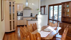 A modern kitchen with wooden cabinets, a dining table with chairs, and a large window providing natural light. - Kurrajong Views Jindabyne (Jindabyne)