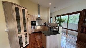A modern kitchen with wooden cabinets, a central island, and a large window providing a view of the outdoors. - Kurrajong Views Jindabyne (Jindabyne)