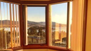 A corner room with a large bay window overlooking a lake and distant hills. There are two dark-colored sofas and a small table with a patterned tablecloth. - Kurrajong Views Jindabyne (Jindabyne)