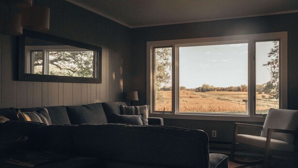 A living room with a dark-colored sofa, a chair, and a large window offering a view of a field. - Cozy remodeled modern farmhouse for up to 10, with secondary room for up to 30 (New London)