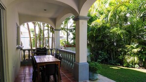 Une terrasse couverte, dotée d’une table et de chaises, entourée d’une végétation luxuriante et recouverte d’un sol carrelé.