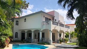 Une maison à deux étages avec une piscine, entourée d’une végétation luxuriante et de palmiers.