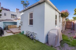 Une maison de plain-pied avec une petite terrasse, un barbecue extérieur abrité et un tuyau d’arrosage raccordé à une source d’eau.