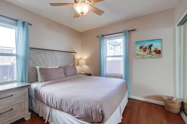 Un dormitorio con una cama, una mesita de noche, un ventilador de techo y una ventana con cortinas.