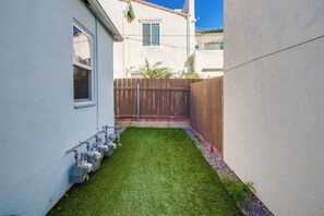 A narrow backyard with artificial grass, a wooden fence, and a utility meter on the wall. - Charming PB bungalow, pet-friendly yard, parking (San Diego)