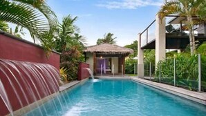 En swimmingpool med et vandfald, omgivet af frodig vegetation og en bindingsværksbygning.