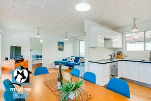 A modern apartment with a living room, dining area, and kitchen. There is a blue sofa, a dining table with chairs, and a kitchen with white cabinets and a stove.