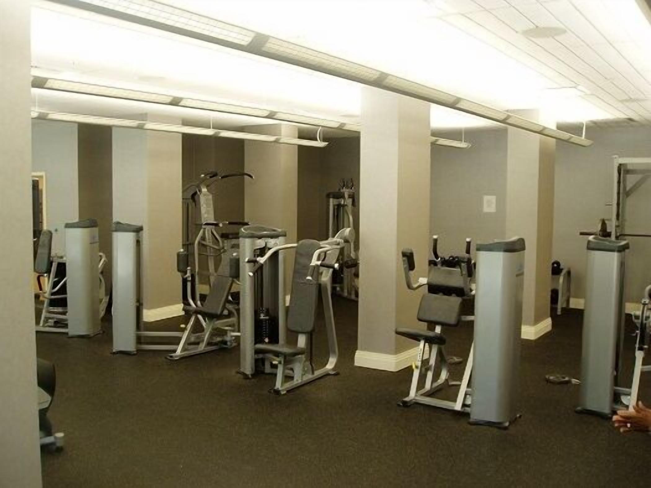 Een sportschool met diverse fitnessapparaten, waaronder elliptische trainers en krachtapparaten, in een ruime kamer met hoge plafonds en fluorescent verlichting.