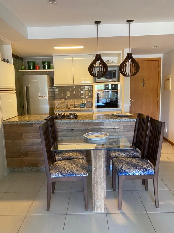 Una cocina moderna con una isla central, dos lámparas colgantes y un comedor con mesa de vidrio y sillas estampadas.