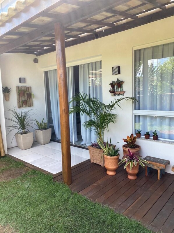A covered patio area with a wooden deck, potted plants, and a window with curtains.