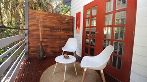 Una terraza de madera con dos sillas blancas y una mesita, una puerta roja con paneles de vidrio, y una cerca de madera.