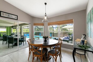 A dining area with a round table and chairs, large windows with blinds, a hanging light fixture, and a view of a pool and outdoor seating. - 5 Minute Walk To Beach & JW Marriott, Fantastic SW Pool Exposure, Designer Home (Marco Island)