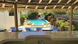 Un espace piscine doté d’un toit de chaume, de chaises longues bleues et d’une piscine en béton entourée de plantes et d’une clôture en bois.