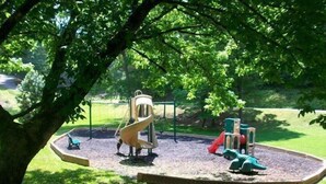 Une aire de jeux avec un toboggan, des structures d’escalade et une balançoire, le tout entouré d’arbres et d’une pelouse.