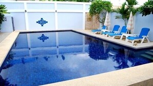 Una piscina rectangular con un diseño de mosaicos, rodeada de sillas de descanso y una pared blanca con un diseño geométrico azul.