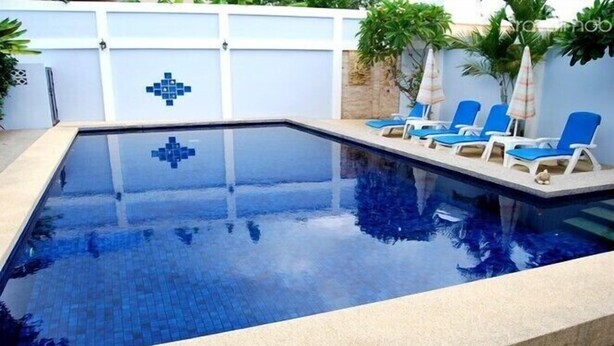 Una piscina rectangular con un diseño de mosaicos, rodeada de sillas de descanso y una pared blanca con un diseño geométrico azul.