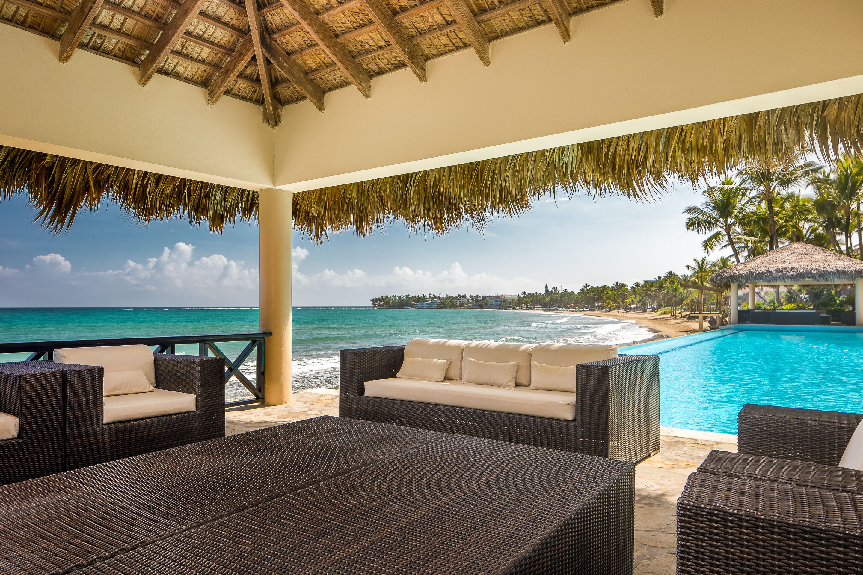A covered outdoor seating area with wicker furniture, a pool, and a view of the ocean and beach.