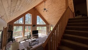 Un amplio salón con techo alto, ventanas grandes, chimenea, televisor y escalera.