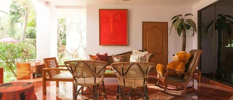 Una habitación con muebles de mimbre, un cuadro abstracto rojo y un ventanal con vistas a la vegetación.