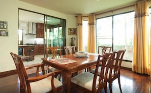 A dining room with a wooden table and chairs, a kitchen in the background, and large windows with curtains.
