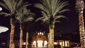 Een nachtzicht van een hotel met palmbomen versierd met lichtjes, een zwembad en een gebouw met bogen.