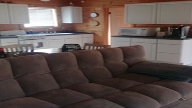 A cozy cabin interior with a sectional sofa, wooden ceiling, white cabinets, and a kitchenette.