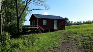 A small, single-story red cabin with a metal roof, a wooden deck, and a door.